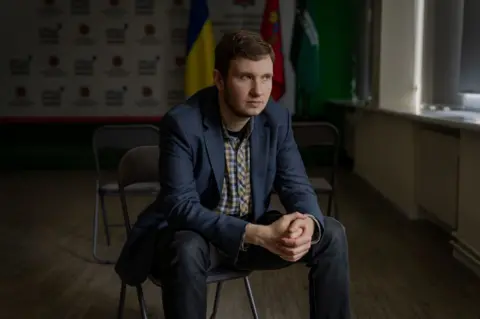 Local prosecutor Mykyta Manevskyi, who is wearing jeans, a blue suit jacket and yellow checked shirt, sits on a chair in a media room in the district prosecutor