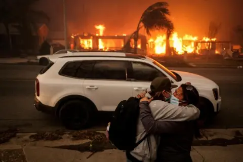 Reuters Two women are visibly upset as they embrace, with fires raging in the background. They are wearing face masks and dark clothes and one of them carries a rucsac. A white car stands behind them. The fires have already largely destroyed the buildings on the other side of the road and damaged trees sway in the wind.